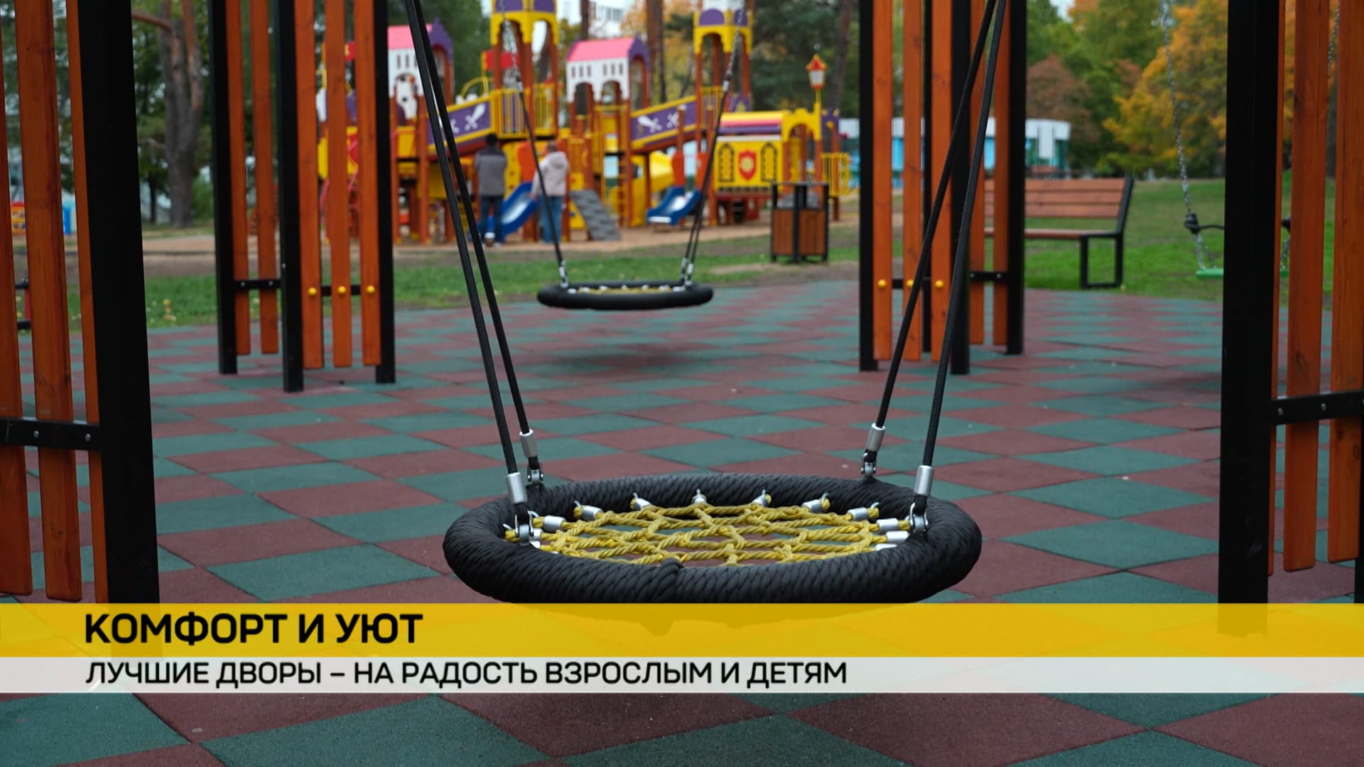 детские качели‑гнездо на площадке: круглая сетчатая конструкция с жёлтой оплёткой, подвешена на чёрных тросах. На фоне — игровая зона в стиле замка, скамейка, деревья