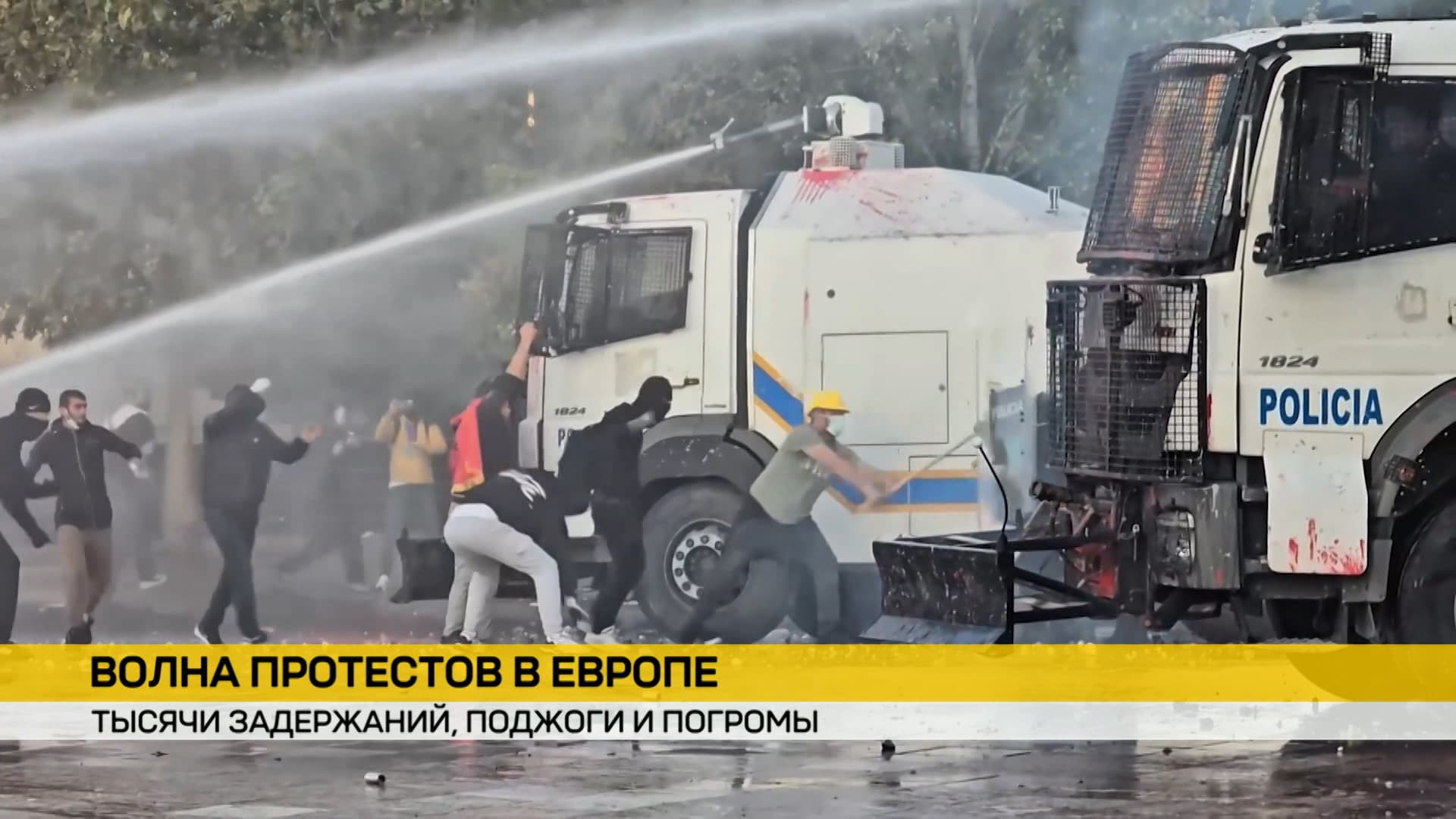 Протестующие в масках атакуют полицейские грузовики с надписью «POLICIA». Спецтехника применяет водометы для разгона толпы среди дыма.